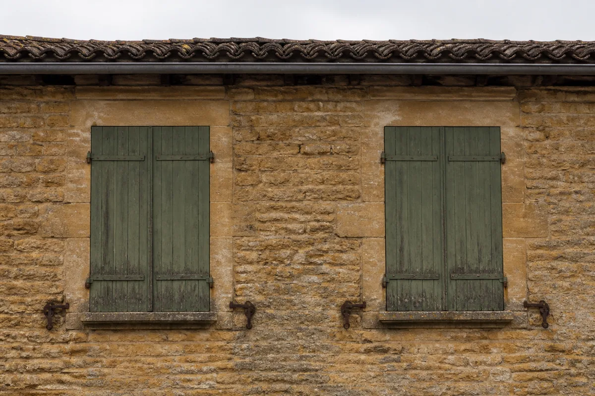 Façade de maison en pierre dorée d’un bourg des Monts du Lyonnais sous un ciel d’hiver bas, volets de bois fermés